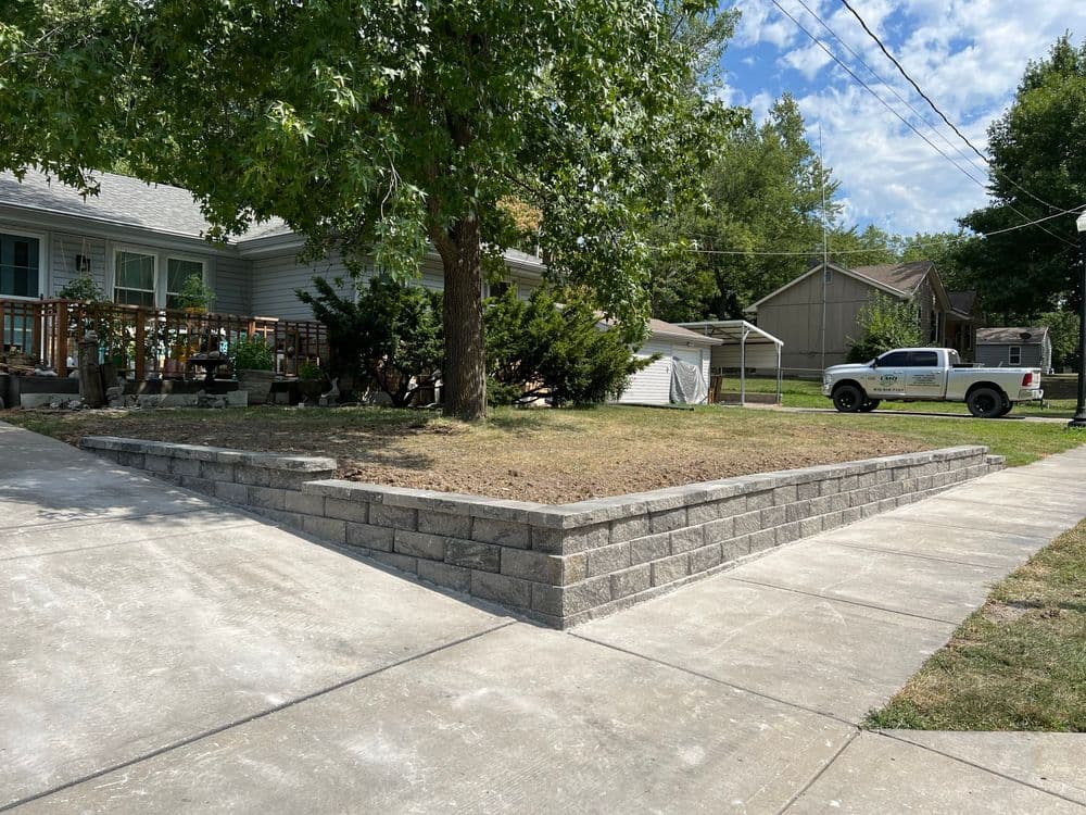Raised stone garden bed with grass, surrounded by driveway and trees in residential area.