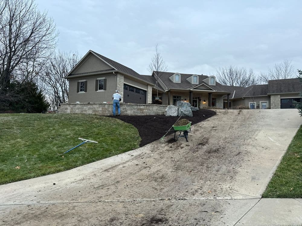 Man landscaping a home yard with mulch and a wheelbarrow on a cloudy day.